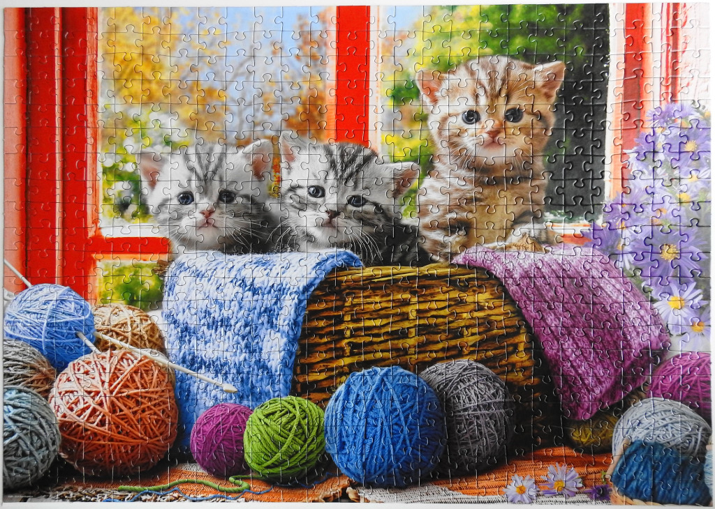 Three kittens sitting in a woven basket surrounded by colorful balls of yarn in front of a window.