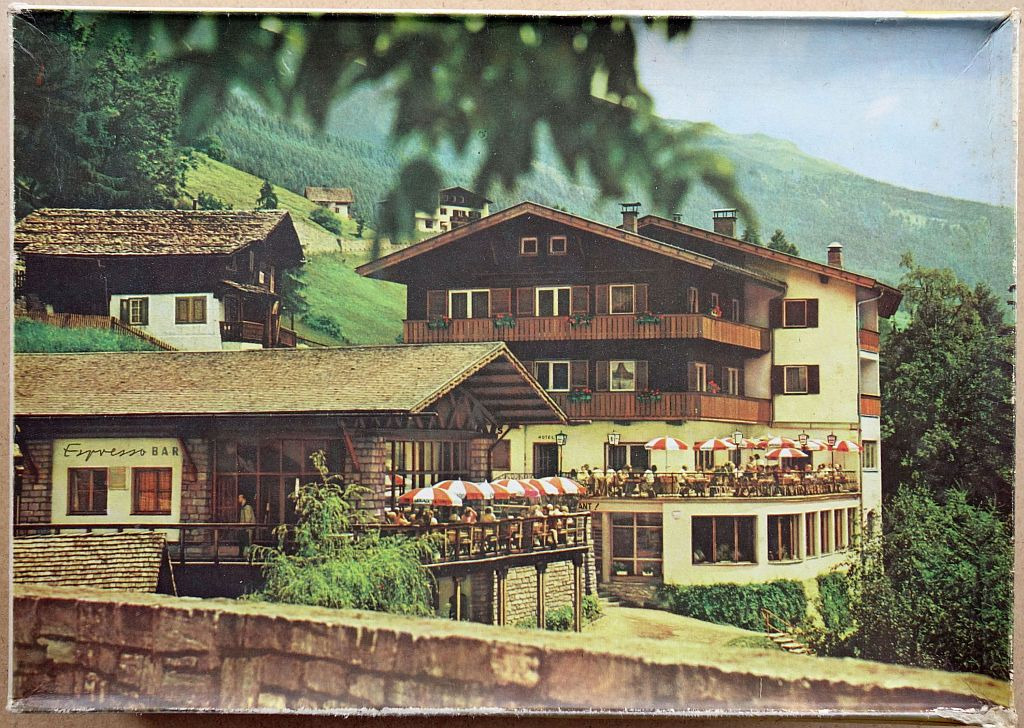 A vintage photograph of an alpine hotel with an outdoor terrace and an Espresso Bar nestled in a green mountain valley.