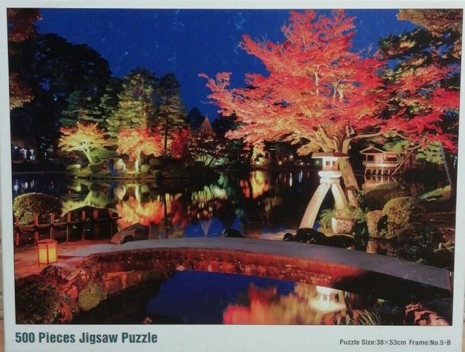 Night view of a Japanese garden with illuminated red autumn maple trees reflected in a calm pond over a stone bridge.
