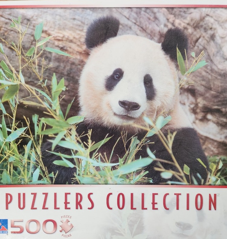A close-up photograph of a giant panda looking forward, partially obscured by green bamboo leaves.