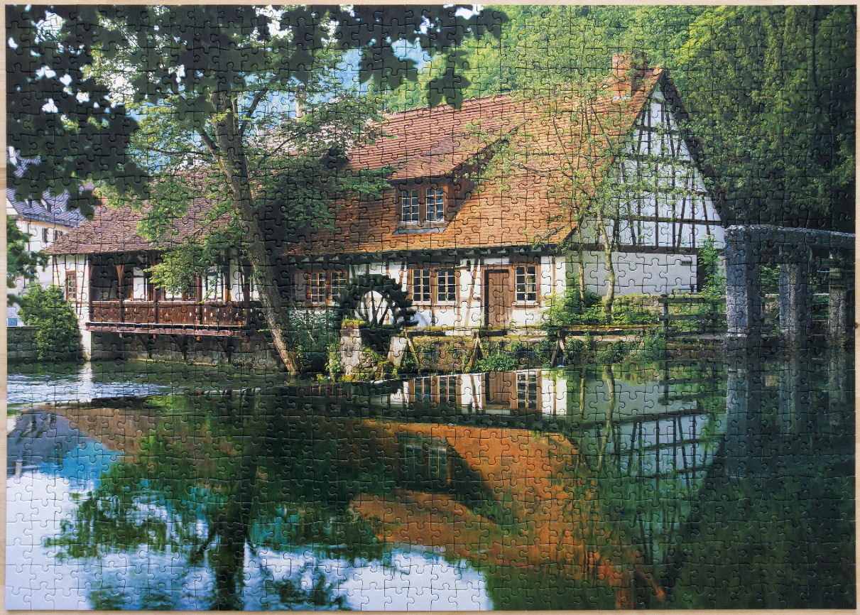 A scenic photograph of a rustic half-timbered water mill reflecting in a still, clear pond.