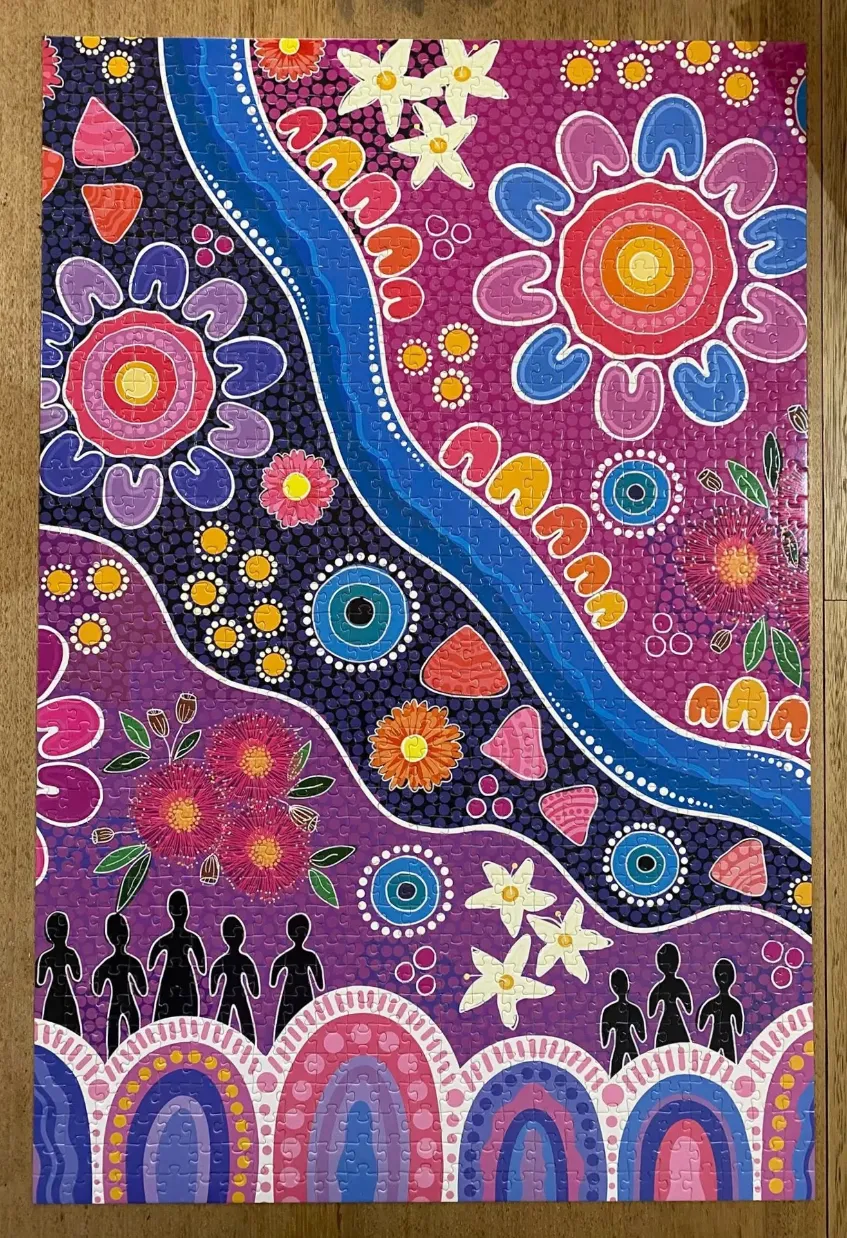 A vibrant Indigenous-style dot painting featuring a blue river, stylized flowers, and silhouettes of people.