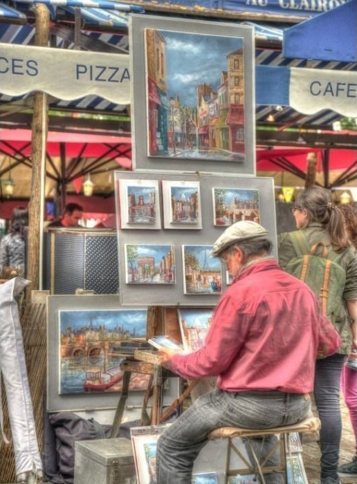 An artist in a pink shirt sits on a stool painting at a street market in Paris.