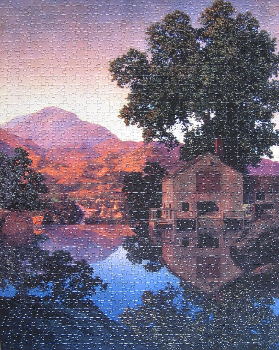A rustic cottage sits on a calm lake reflecting the pink and orange sunset over distant mountains.