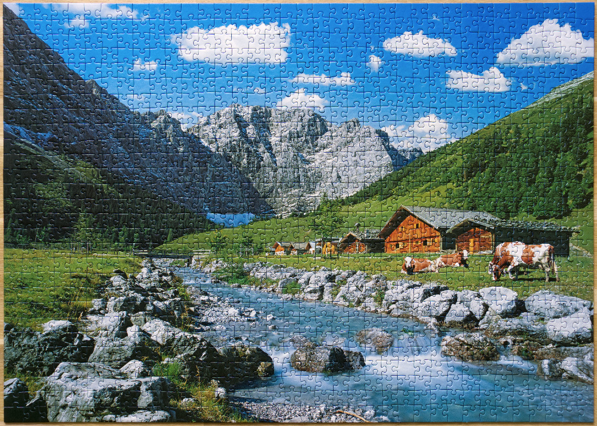 A sunny mountain valley landscape with a rocky stream, wooden cabins, and cows grazing in an alpine pasture.