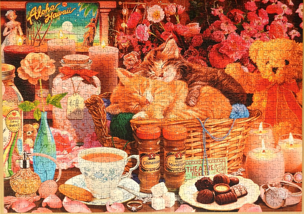 Two kittens sleeping in a wicker basket surrounded by flowers, candles, tea, and chocolates.