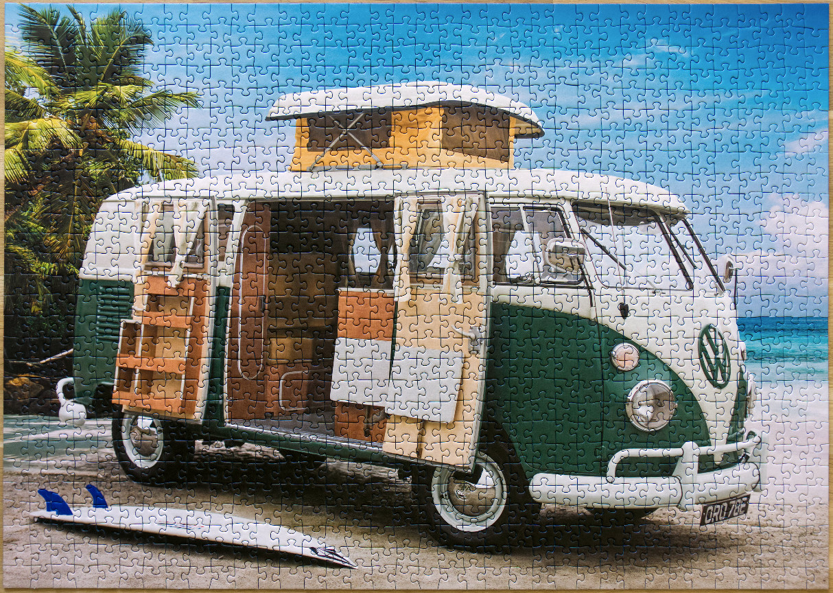 A vintage green and white Volkswagen camper van parked on a sandy beach next to a surfboard with palm trees in the background