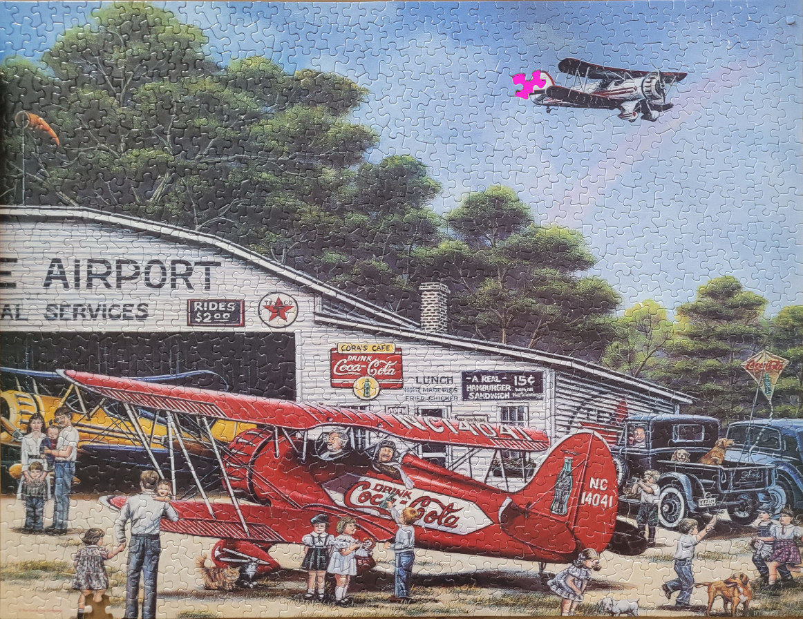 A vintage, red Coca-Cola biplane parked outside a country airport hangar with people and classic cars.