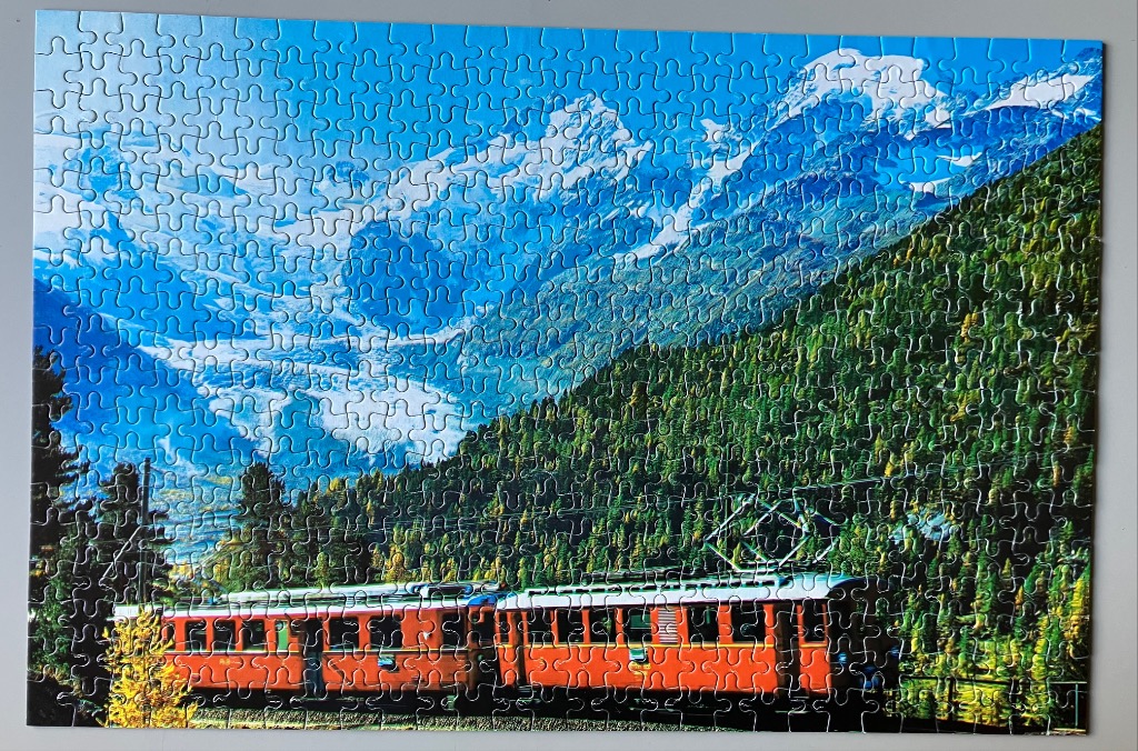 A bright red mountain train traveling through a lush green alpine forest with snowy peaks in the background.