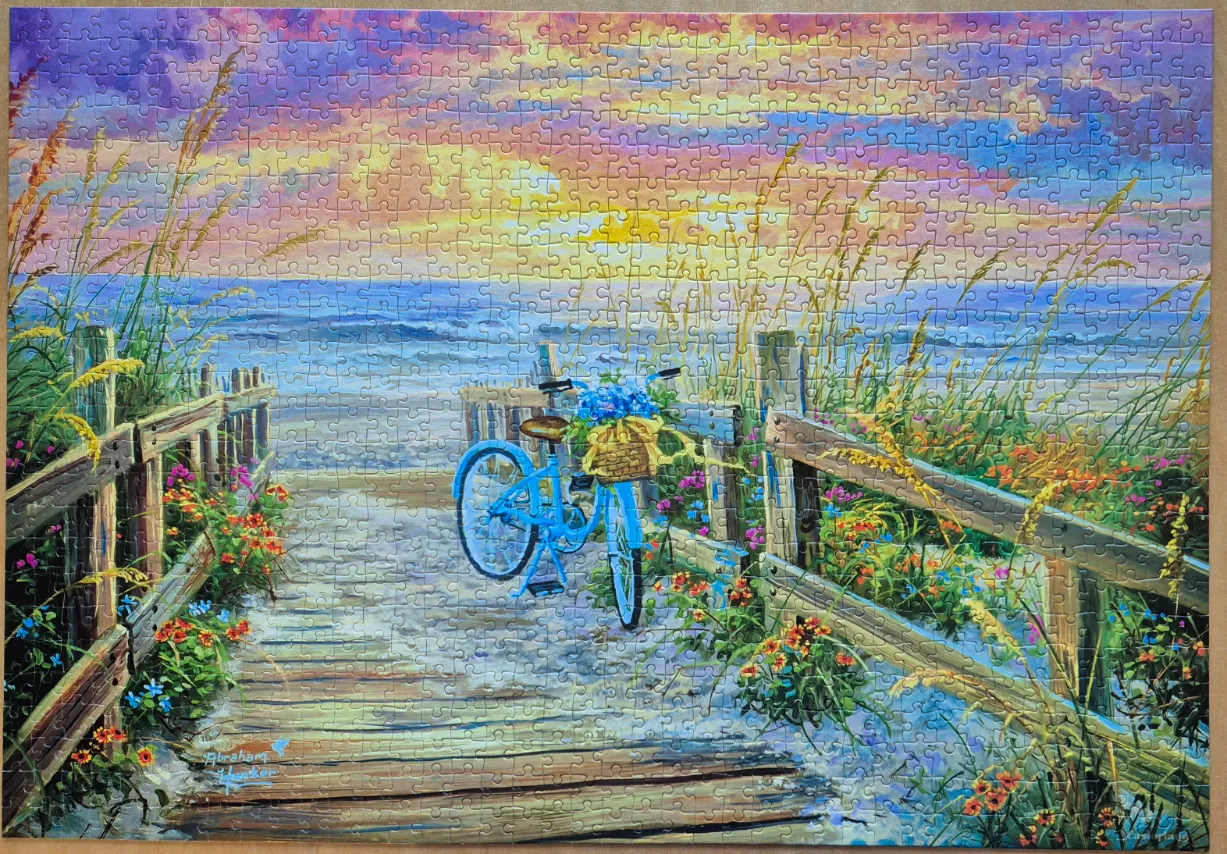 A blue bicycle with a flower basket parked on a wooden boardwalk leading to a sunset beach scene.