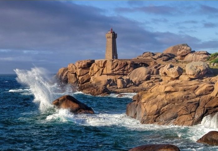 A stone lighthouse standing on rugged, pink-hued granite rocks against a sea with crashing waves.