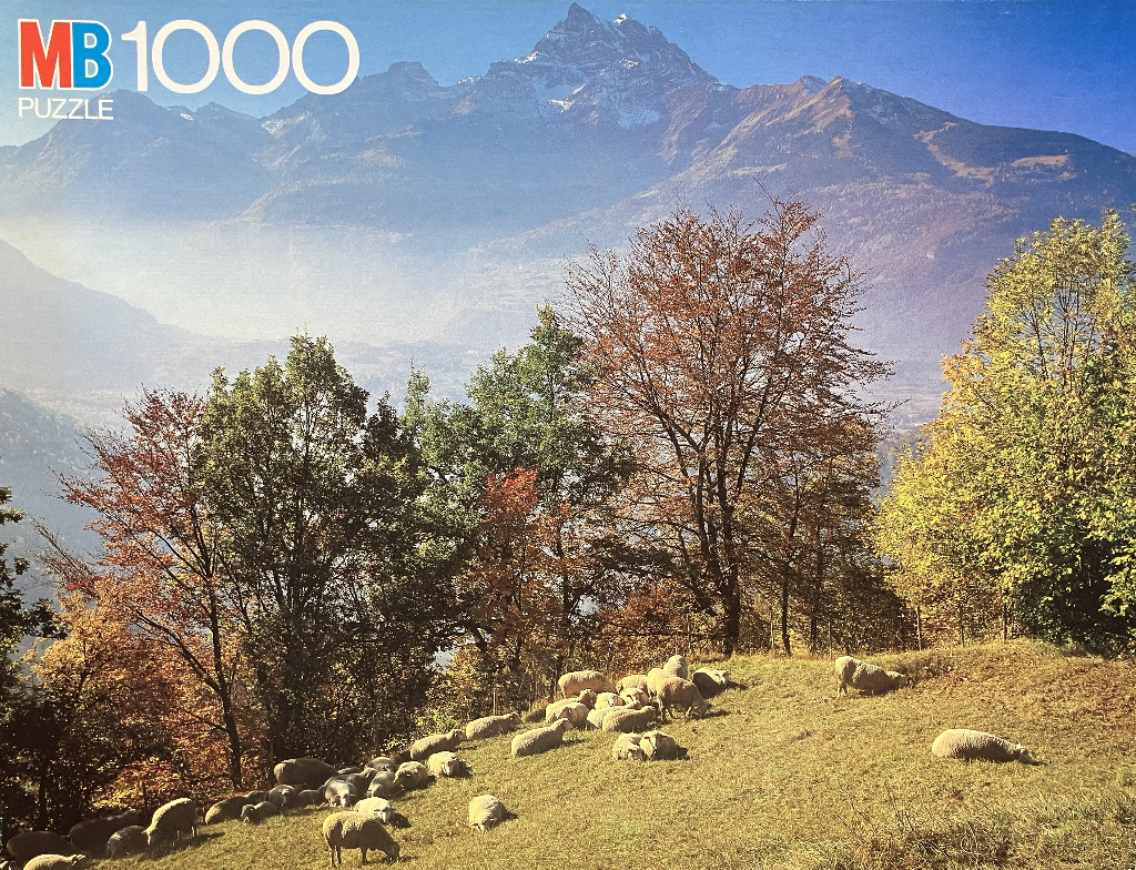 A flock of sheep grazing on a hillside meadow beneath tall, autumn-toned trees and distant snowy mountains.