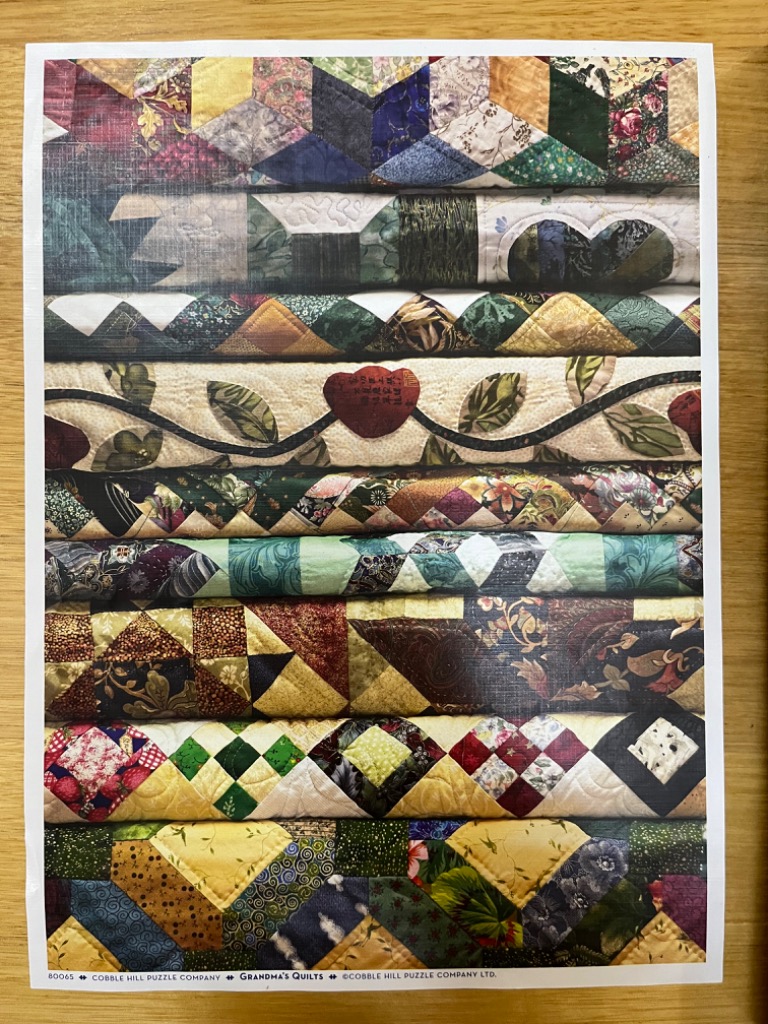 A stack of nine folded, colorful, patterned fabric quilts.
