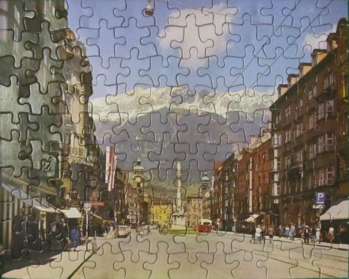 A vintage photograph of a European city street lined with buildings and framed by snow-capped mountains.