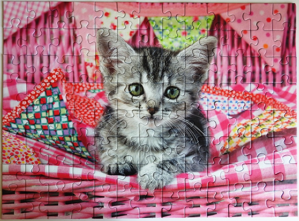 A grey tabby kitten sitting in a pink wicker basket filled with colorful patterned fabrics.