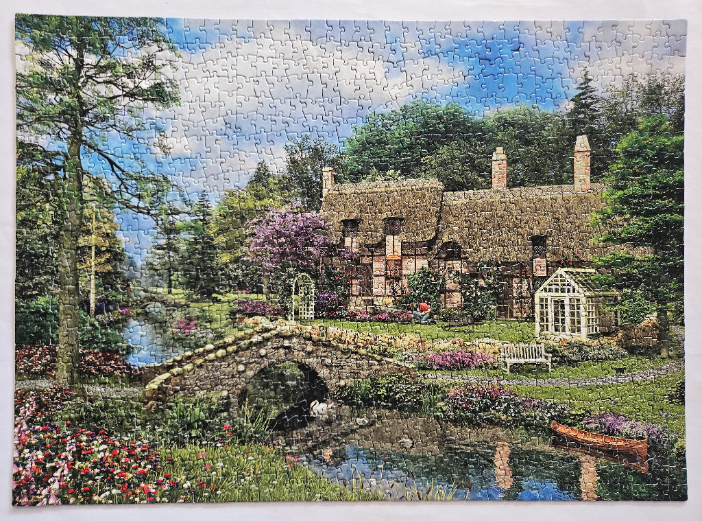 A charming thatched-roof cottage nestled beside a stone bridge and a tranquil pond in a lush garden.