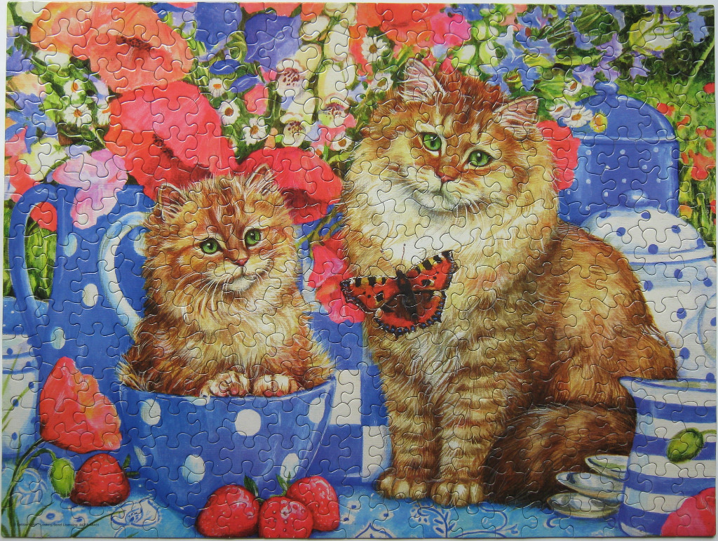 A fluffy ginger kitten sits in a blue polka-dot tea cup beside a larger ginger cat with a butterfly on its chest.