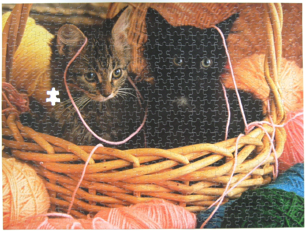 Two kittens, one tabby and one black, sitting inside a woven basket surrounded by balls of yarn.