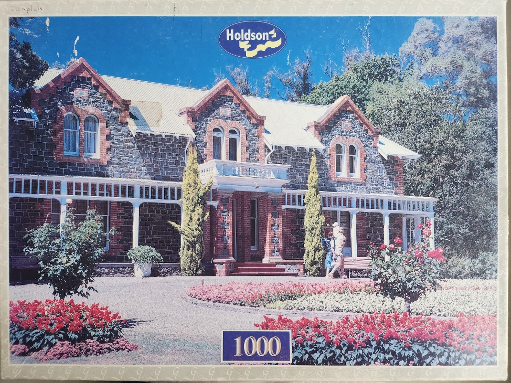 A stately stone country manor house with a manicured garden and red floral beds in the foreground.