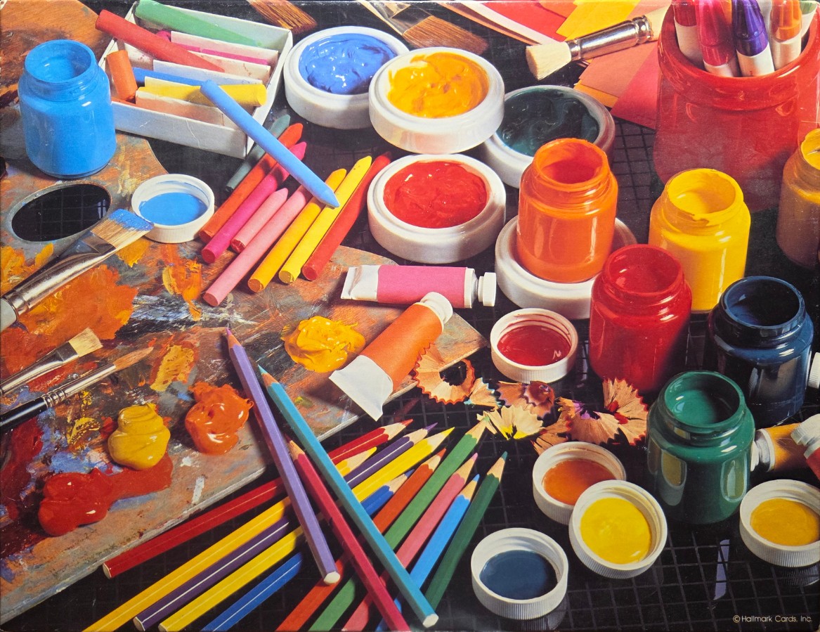 An assortment of vibrant art supplies, including paint pots, tubes, crayons, and colored pencils on a dark grid surface.