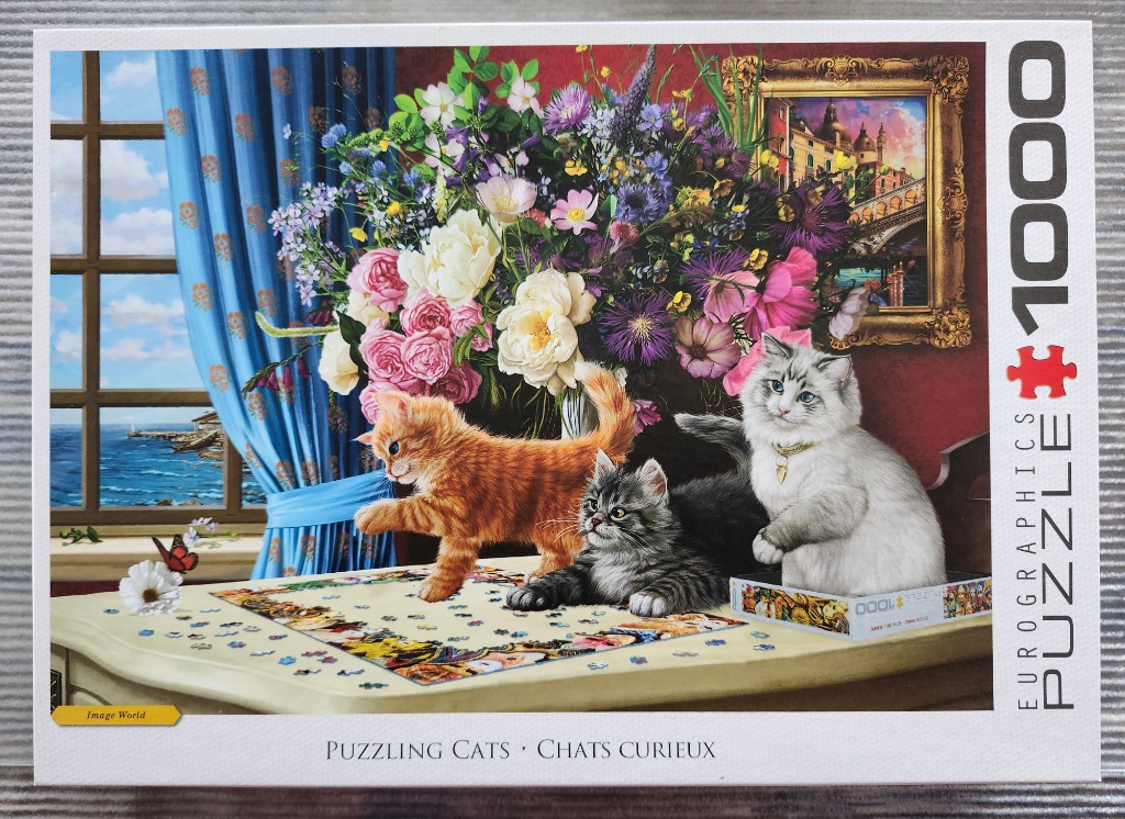 Three kittens sitting on a table with a jigsaw puzzle, near a bouquet of flowers and a window overlooking the ocean.