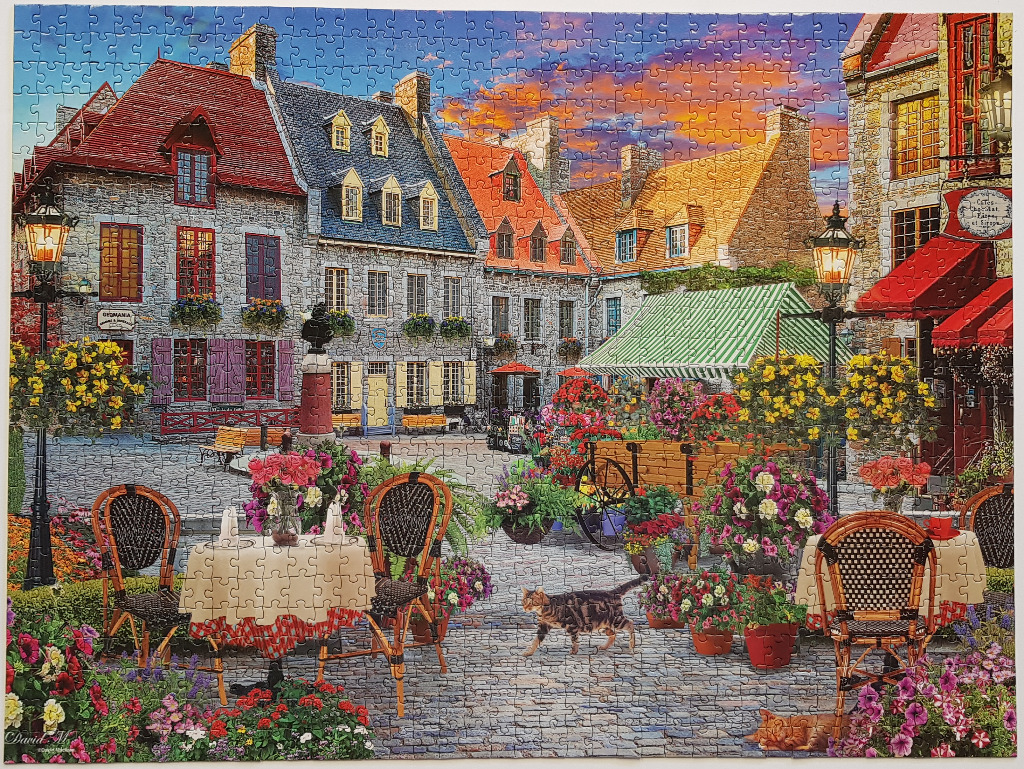 An illustration of a quaint European cobblestone square at sunset with outdoor cafe seating and flower-filled planters.