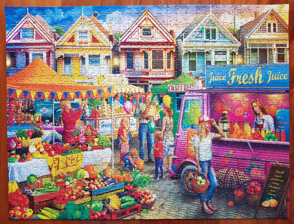 A bustling outdoor farmers market scene set in front of colorful Victorian-style houses.