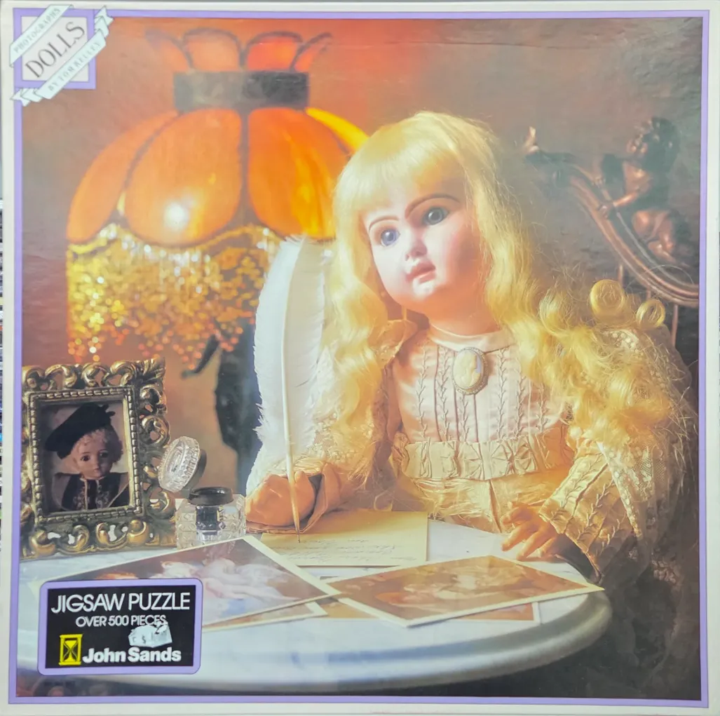 A blonde vintage-style doll writing with a quill at a desk illuminated by a warm orange lamp.