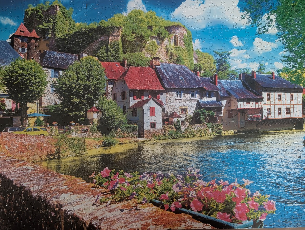 A picturesque European riverside village featuring stone buildings with red roofs, a ruined castle above, and pink flowers.