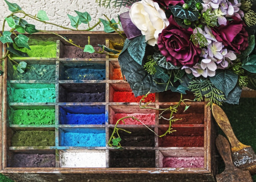 A wooden printer's tray filled with powdered pigments in various colors, accented by a floral arrangement and paint brushes.