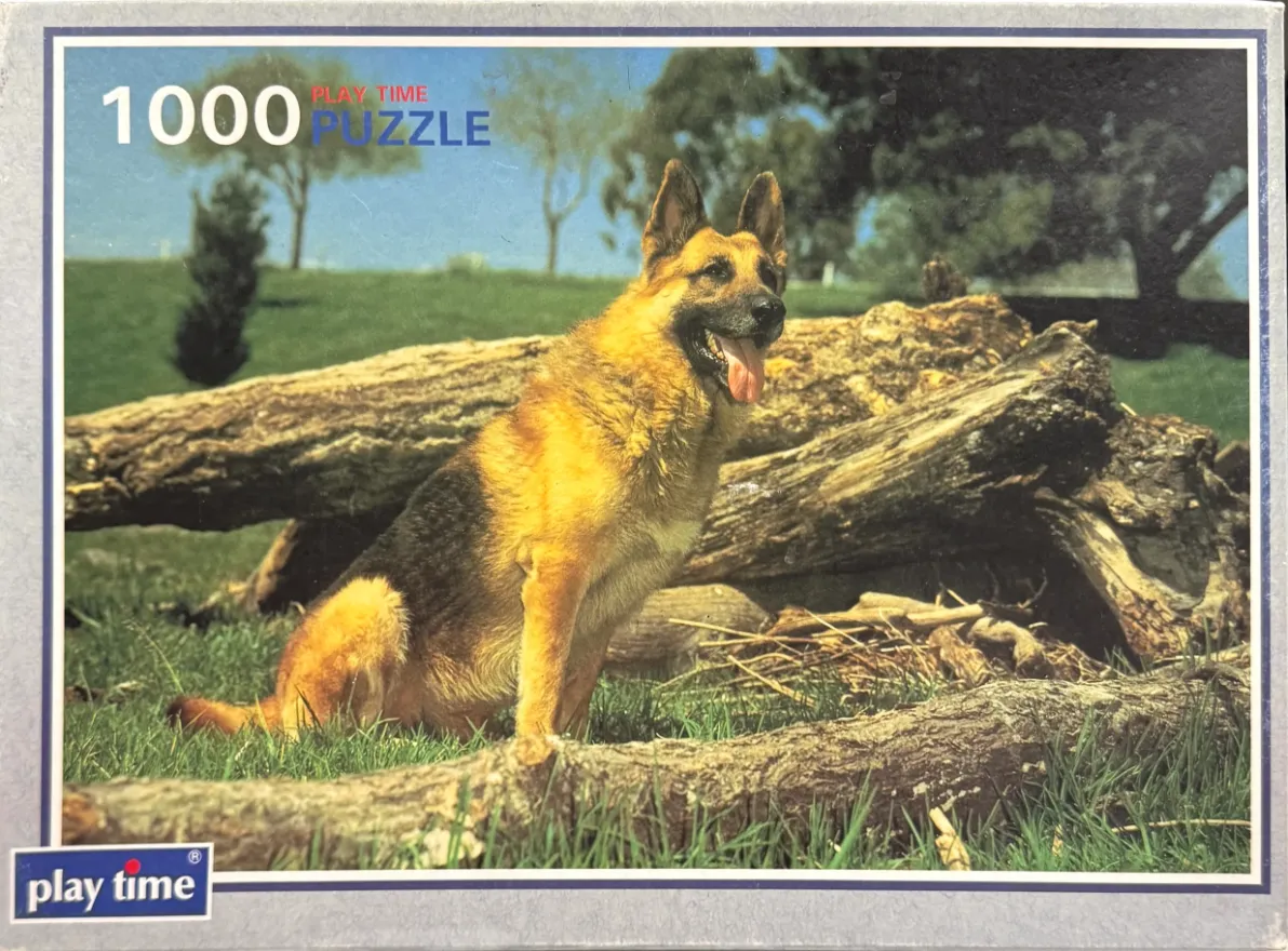 A German Shepherd dog sitting alertly in front of large wooden logs in a grassy outdoor setting.