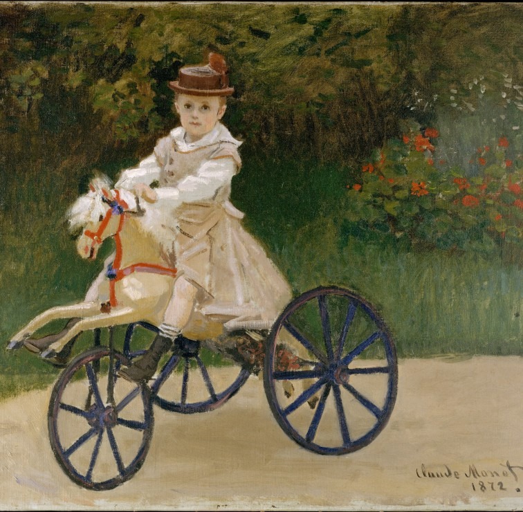 A young child with a hat sits on a wheeled toy horse in a garden.