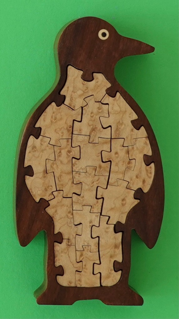 A wooden cutout of a penguin with a dark mahogany frame and a light tan burl wood belly, set against a green background.