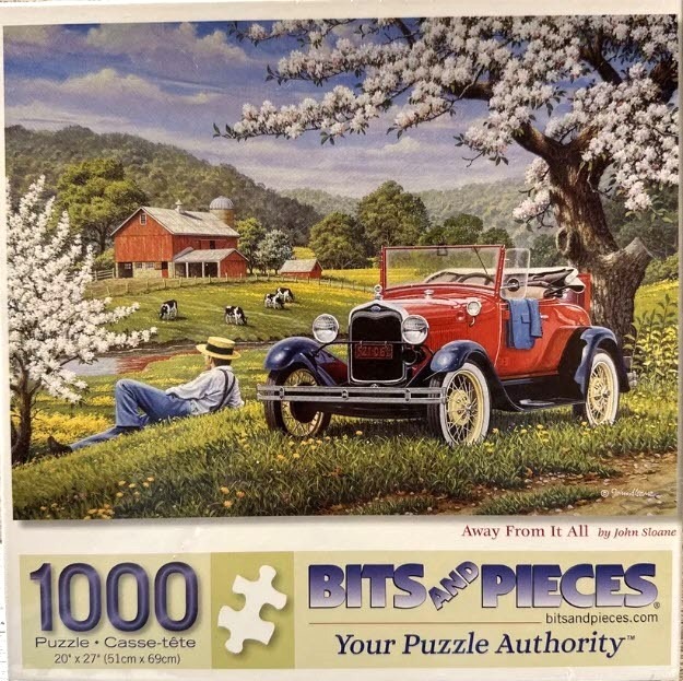 A man in a straw hat relaxes in a field near a red antique roadster with a farm and blossoming trees in the background.
