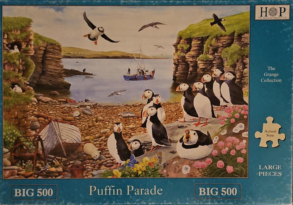 A group of Atlantic puffins on a rocky beach near towering coastal cliffs, with a small fishing boat in the bay.
