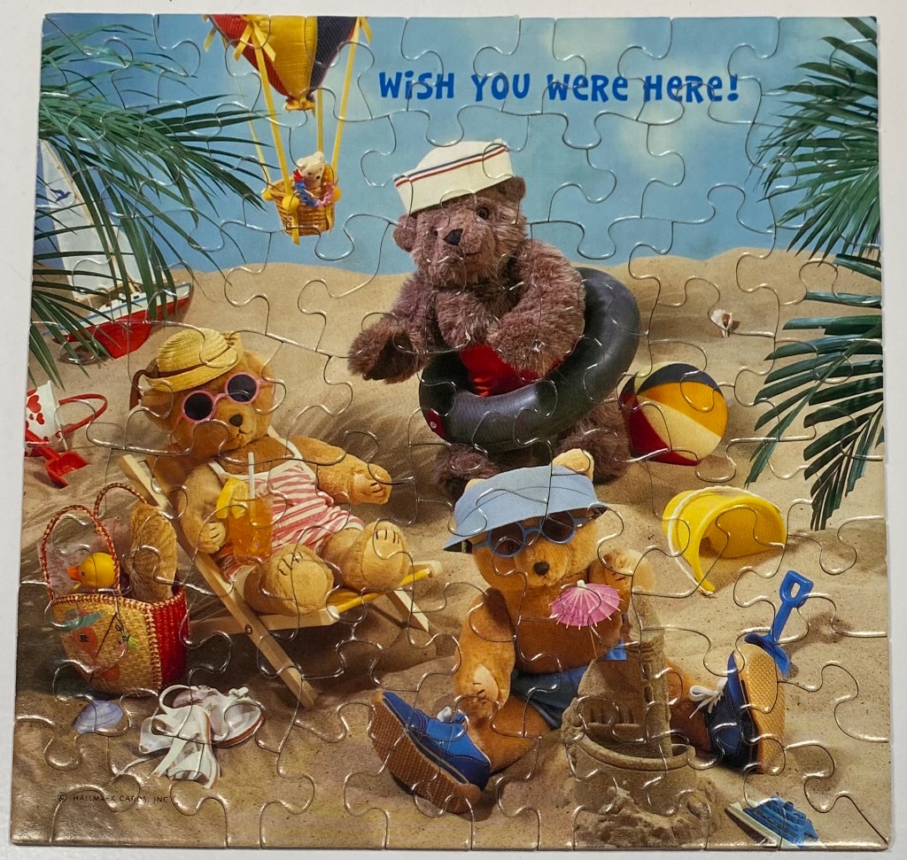 Three teddy bears relaxing on a sandy beach under palm trees, with the text Wish You Were Here! above them.
