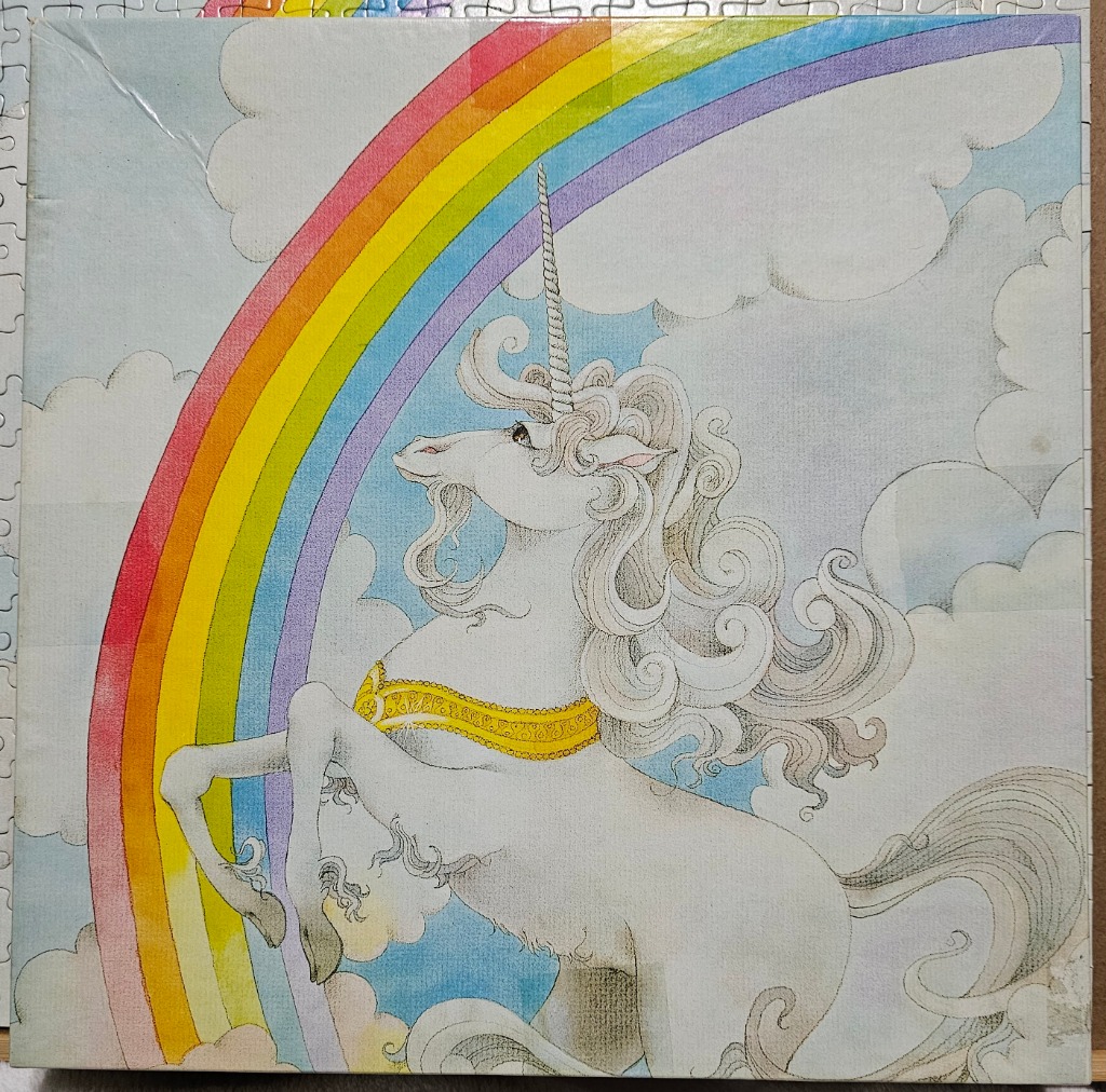 A graceful white unicorn with a gold collar rearing up in front of a vibrant rainbow against a sky of soft clouds.