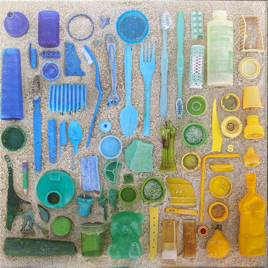 Knolling arrangement of assorted plastic marine debris on sand, graded by color from blue to yellow.