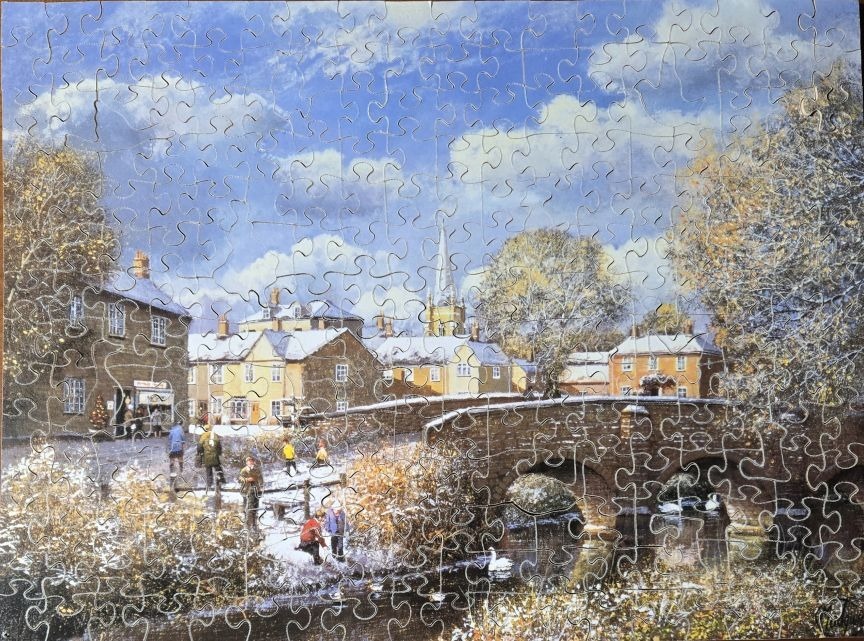 A wintry English village scene with a stone bridge, people walking, and a church spire under a blue sky.