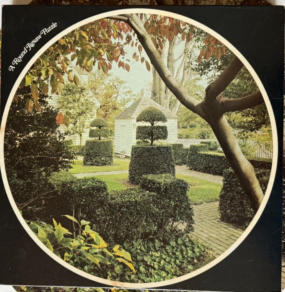 A round garden scene featuring manicured topiary shrubs and brick pathways in front of a white colonial-style building.