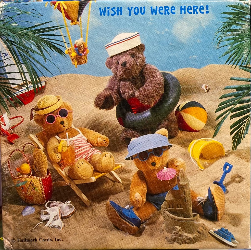 Three teddy bears on a sandy beach under a bright blue sky with a hot air balloon above.