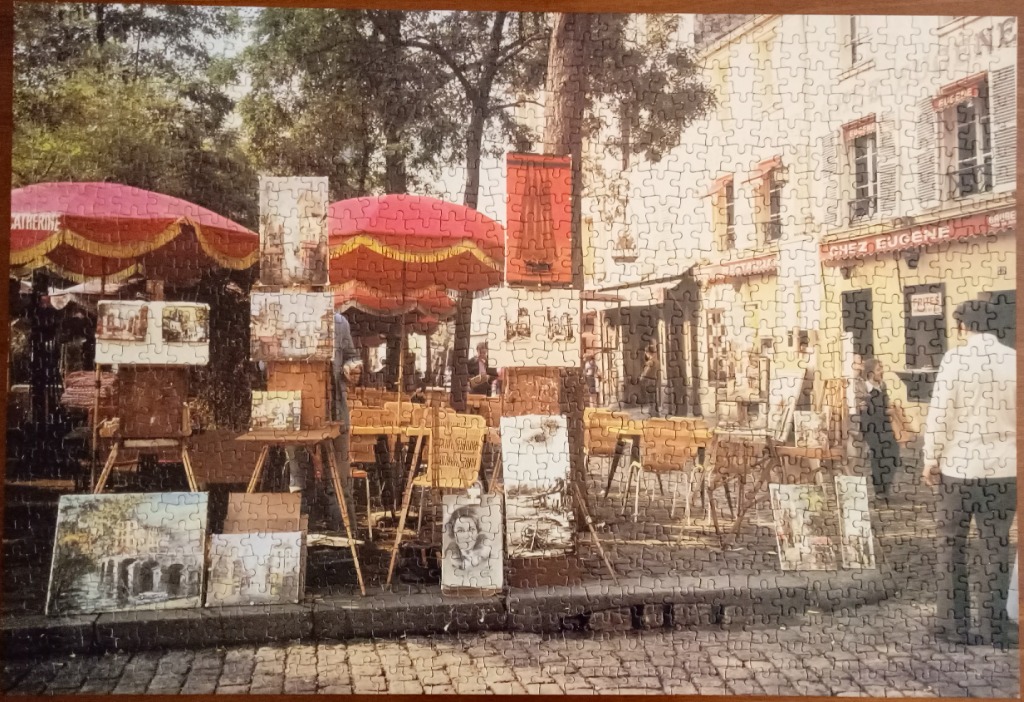An outdoor art market in a Parisian-style town square with red umbrellas and paintings on display.