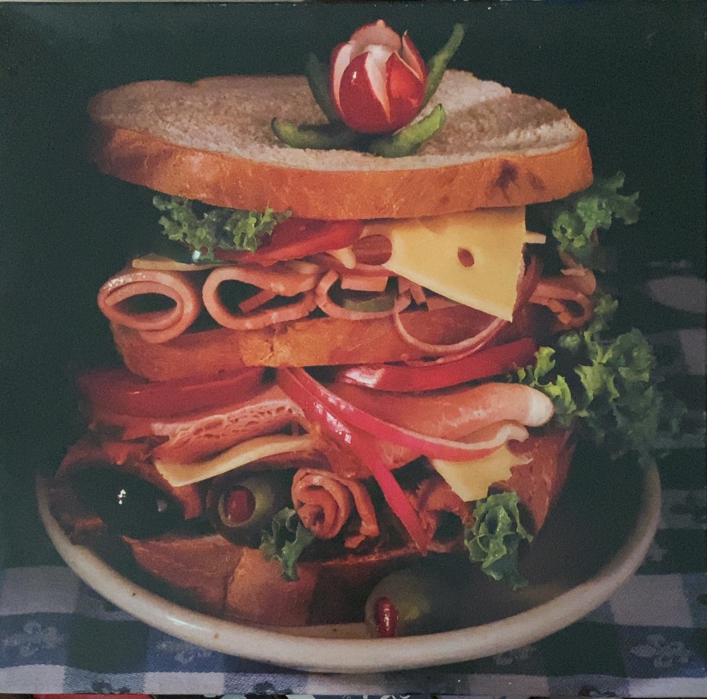 A double-decker sandwich stacked with meat, cheese, lettuce, and a radish garnish on a checked tablecloth.