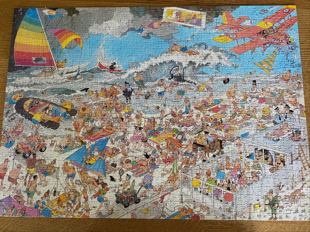 A crowded beach scene featuring a chaotic mix of sunbathers, swimmers, aircraft, and humorous incidents.