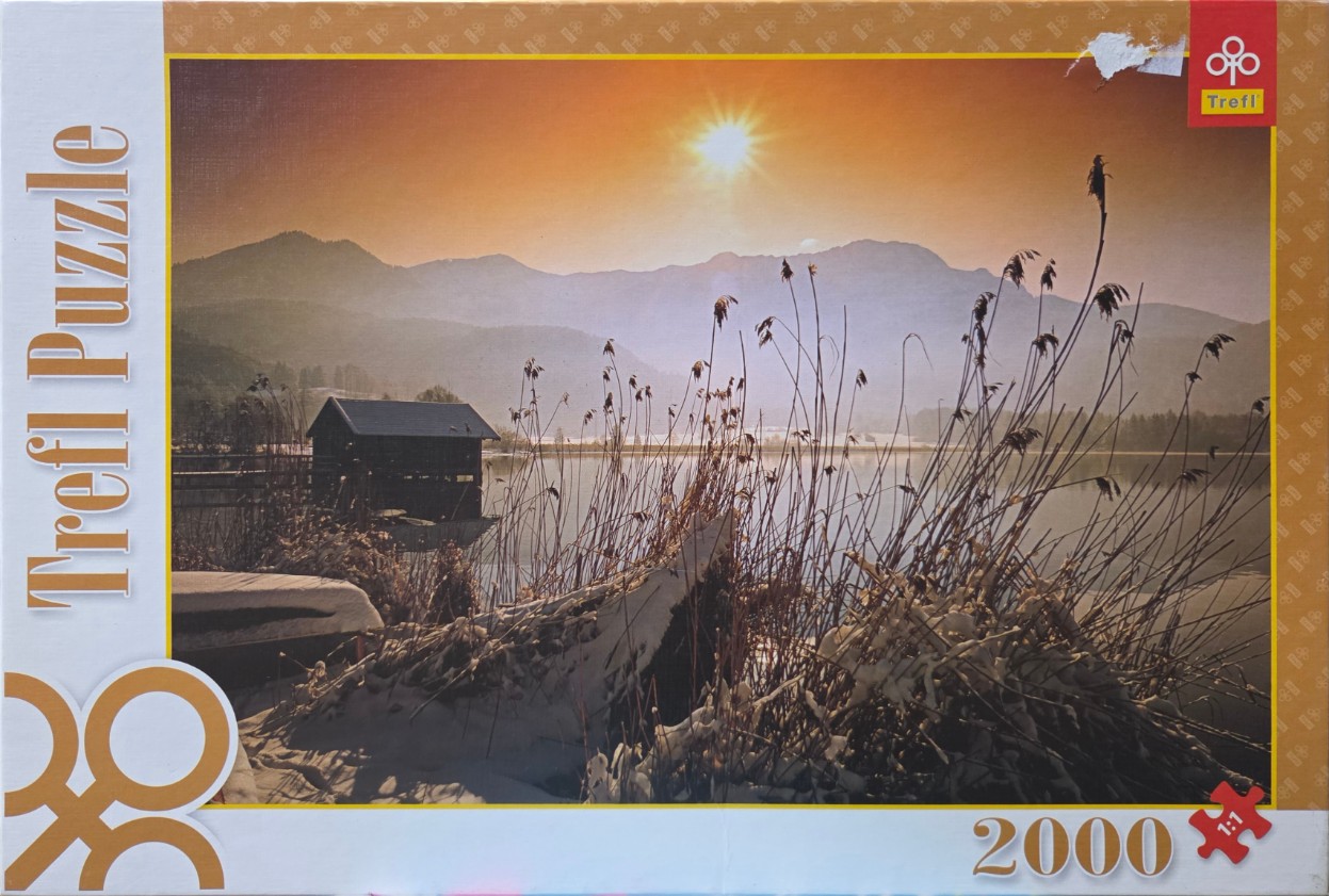 A scenic photograph of a sunlit winter lake with a small cabin and snow-covered reeds.