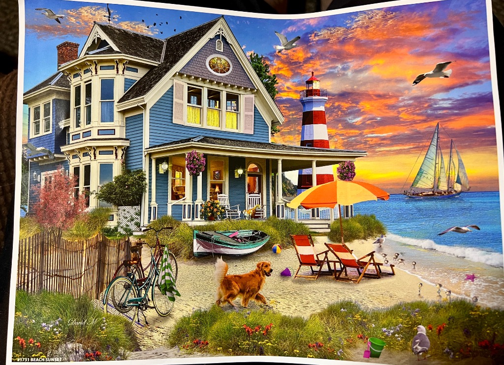 A blue Victorian house on a beach with a lighthouse, sailboat, and dog at sunset.