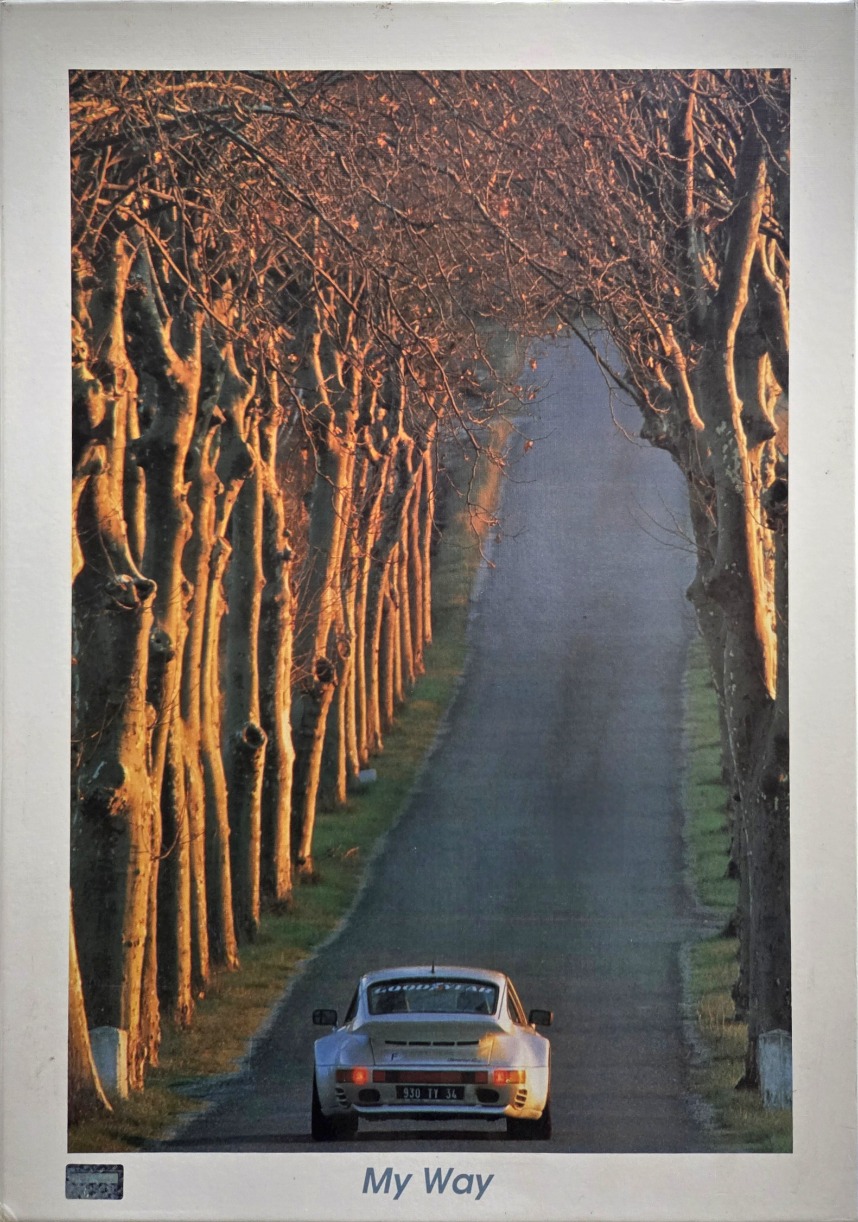 A silver vintage sports car driving down a tree-lined road at sunset.