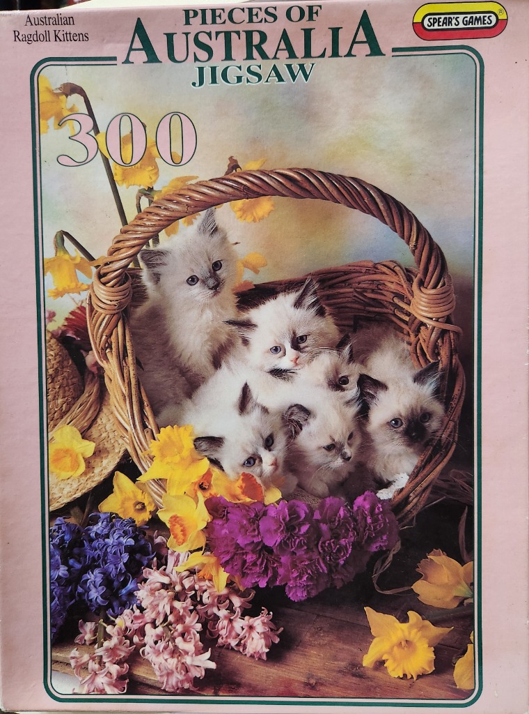 Five fluffy Ragdoll kittens nestled together inside a woven wicker basket surrounded by spring flowers.