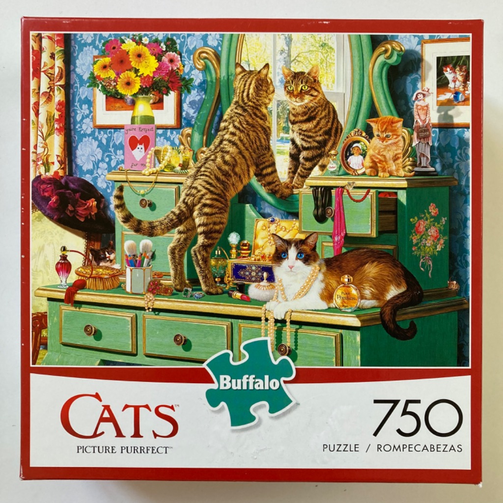 Three cats on a green antique vanity table with perfume, jewelry, and flowers against blue floral wallpaper.
