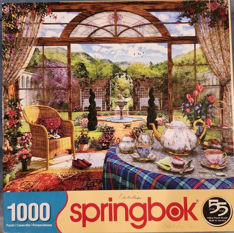 Sunroom overlooking a lush garden with a tea set, wicker chair, and stone fountain.